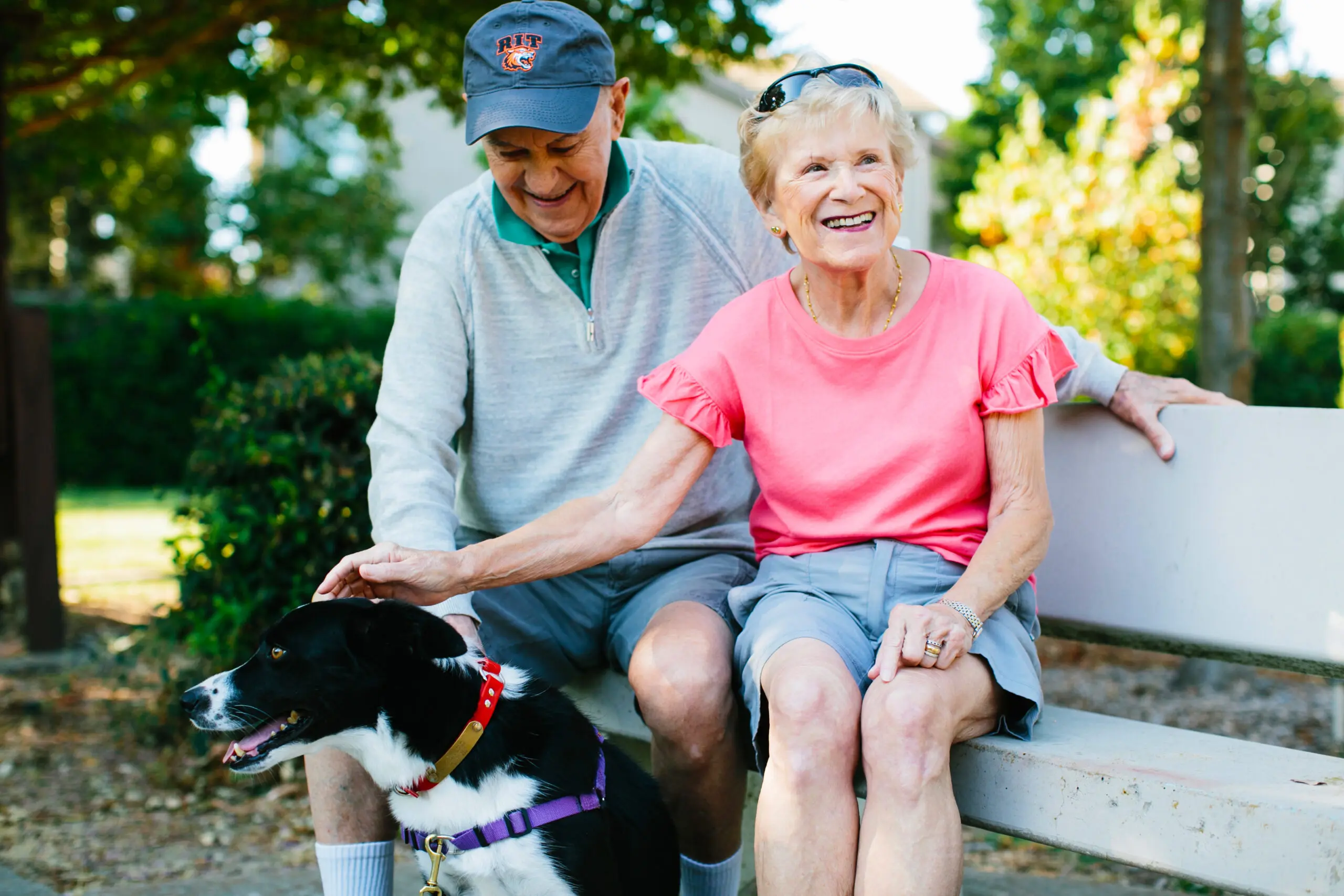 An older couple smiles as they sit on a bench and pet a dog