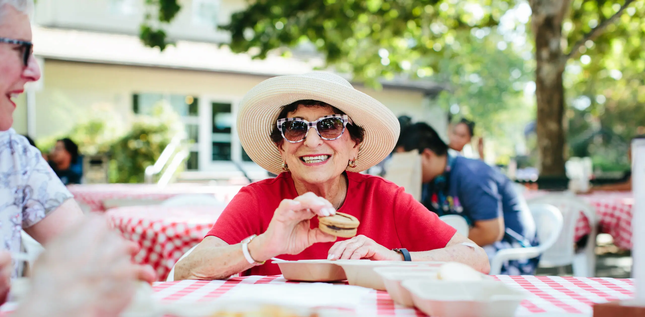 A smiling older woman wearing sunglasses and a hat eats a snack at an outdoor picnic table
